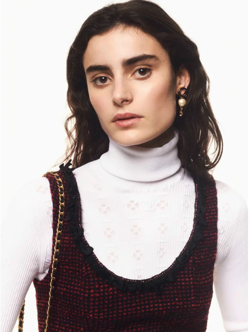 Model wearing a white turtleneck and textured tweed dress with pearl earrings against a solid white background