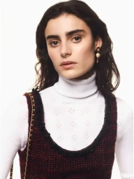 Model wearing a white turtleneck and textured tweed dress with pearl earrings against a solid white background