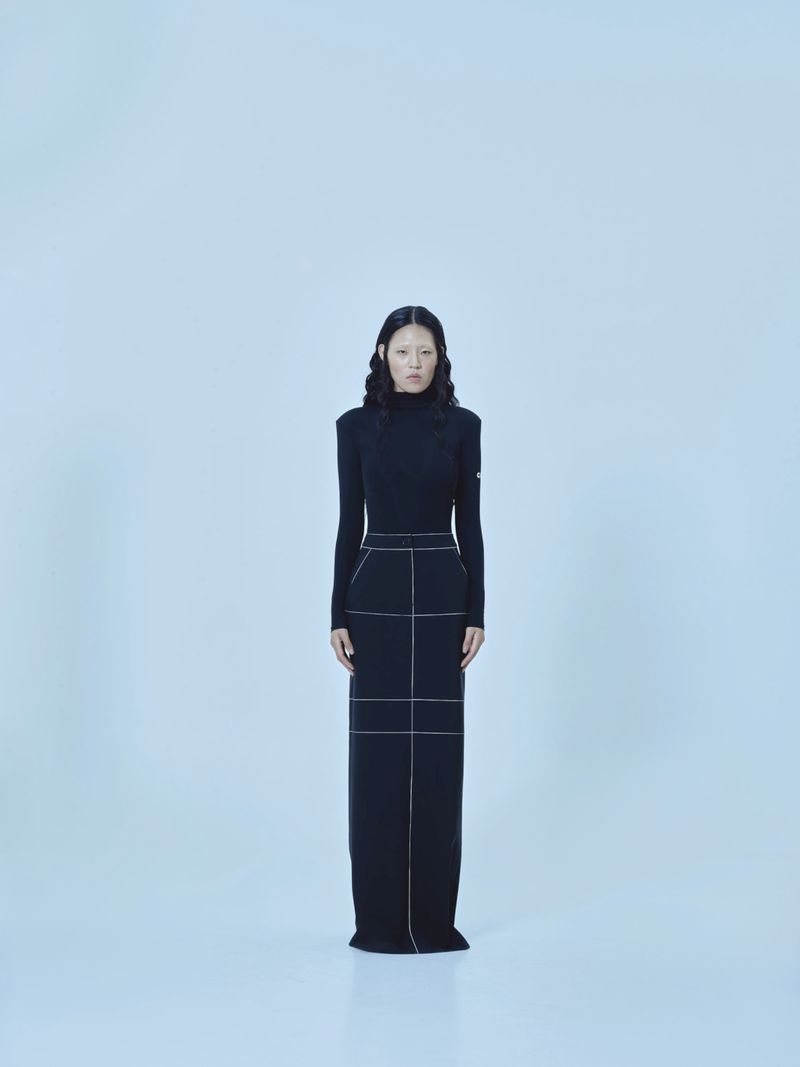 A full shot of an Asian woman standing in a black turtleneck dress against a white backdrop in a fashion or portrait style photo.
