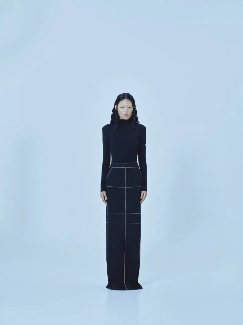 A full shot of an Asian woman standing in a black turtleneck dress against a white backdrop in a fashion or portrait style photo.