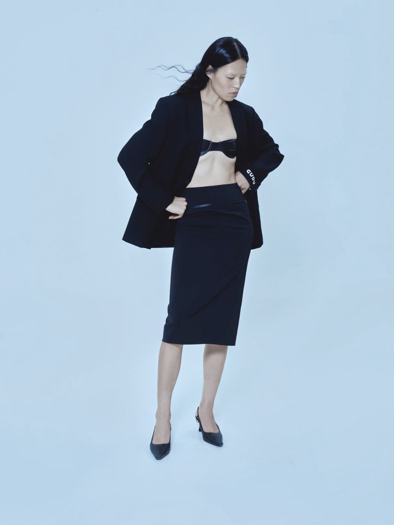 Elegant Asian woman in black blazer, bra, and pencil skirt poses in a fashion studio. Hair is blowing slightly. High fashion, clean aesthetic.