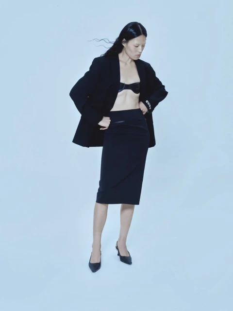 Elegant Asian woman in black blazer, bra, and pencil skirt poses in a fashion studio. Hair is blowing slightly. High fashion, clean aesthetic.