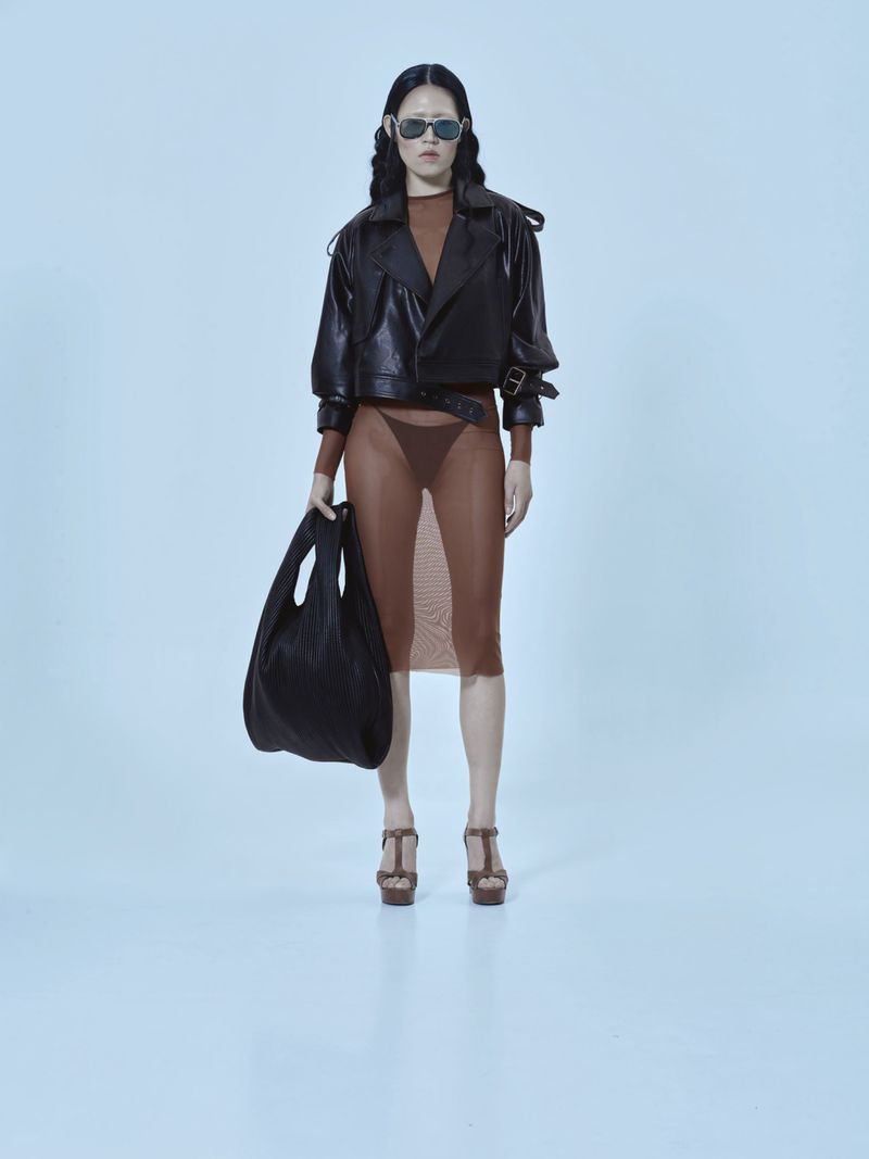 A fashion model strikes a pose in a studio, showcasing a brown mesh dress, black leather jacket, platform heels, and carrying a black tote bag.