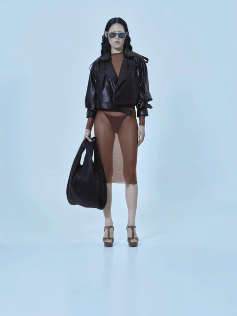 A fashion model strikes a pose in a studio, showcasing a brown mesh dress, black leather jacket, platform heels, and carrying a black tote bag.