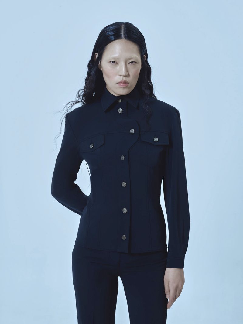 An Asian woman in a black button-down jacket and pants poses against a light blue background, looking directly at the camera in a studio setting.