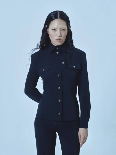 An Asian woman in a black button-down jacket and pants poses against a light blue background, looking directly at the camera in a studio setting.