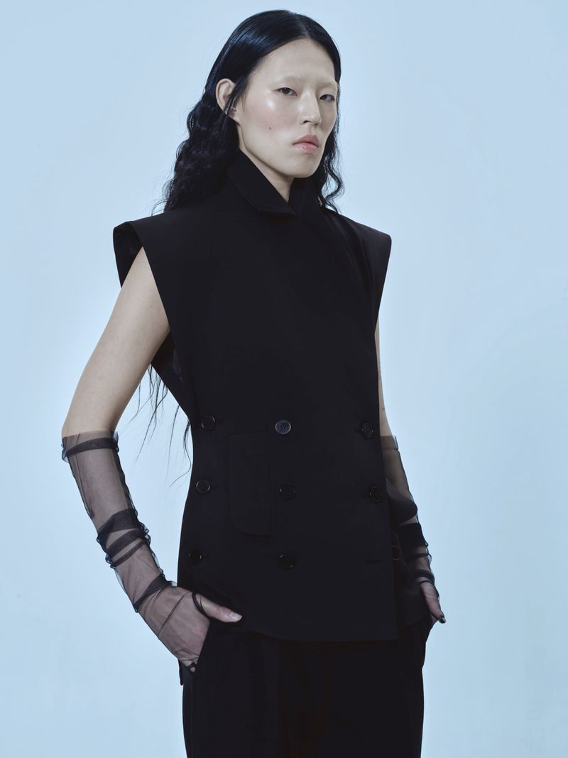 Model in a black sleeveless double-breasted vest and sheer black gloves posing against a pale blue background.
