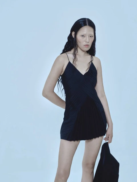 Young adult female fashion model in a black dress with thin straps poses in a studio with a light blue background, holding a garment.
