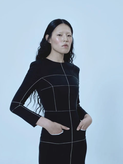 Studio shot of an Asian female model in a black geometric-patterned dress with hands in pockets, against a light blue background.
