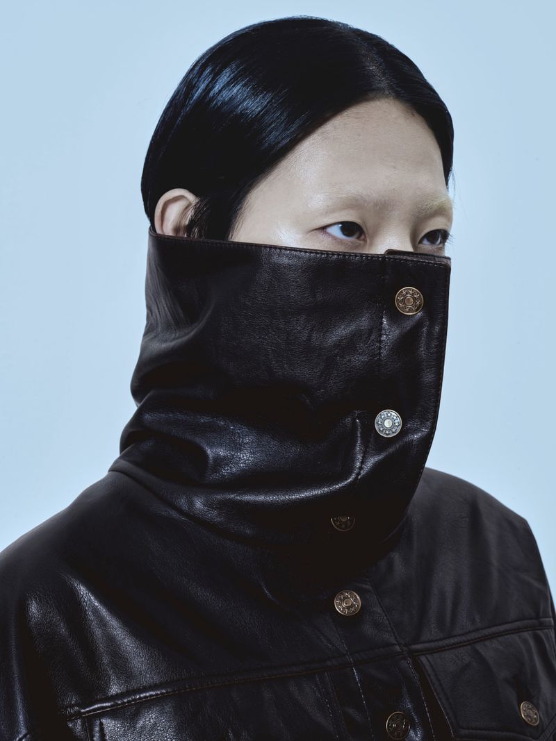 Portrait of a person in a black leather jacket with large collar. The jacket has decorative buttons and is indoors with neutral lighting.