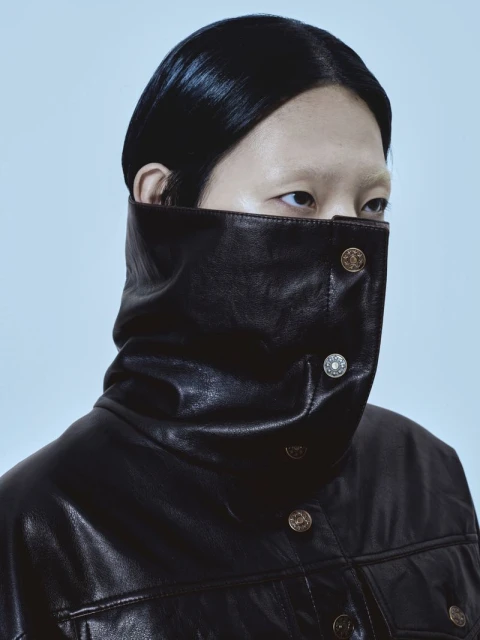 Portrait of a person in a black leather jacket with large collar. The jacket has decorative buttons and is indoors with neutral lighting.