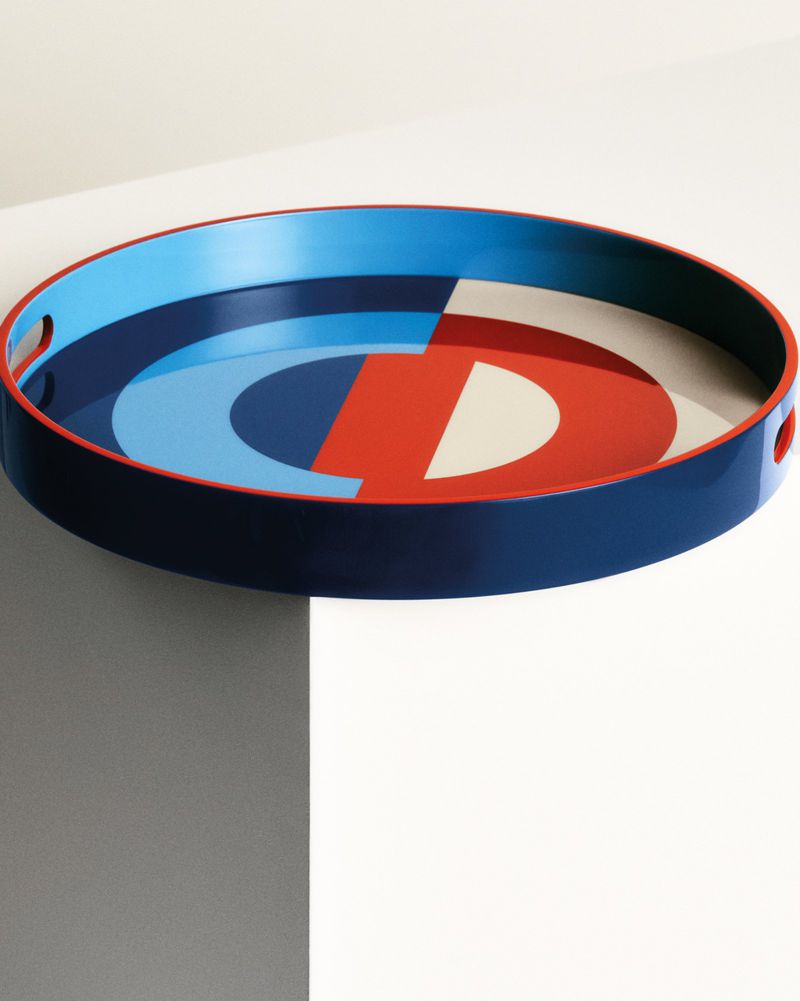A blue-rimmed circular tray with a light blue, dark blue, red, and white geometric pattern sits on a white platform in a studio shot.