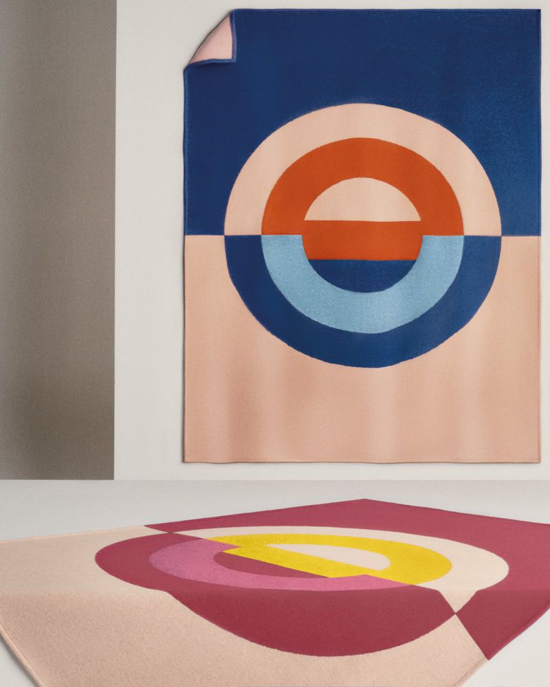 Two blankets with geometric patterns in pink, blue, orange, yellow, and red tones, one draped on a wall and one on a surface.