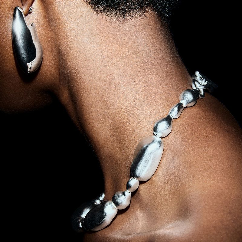Close-up studio shot of a person with dark hair and skin wearing statement jewelry with metallic silver-tone finish against a black background.
