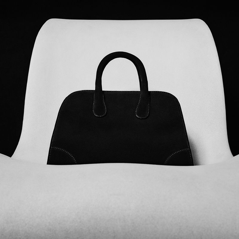 Black structured handbag with top handles resting on a curved white chair against a dark background.