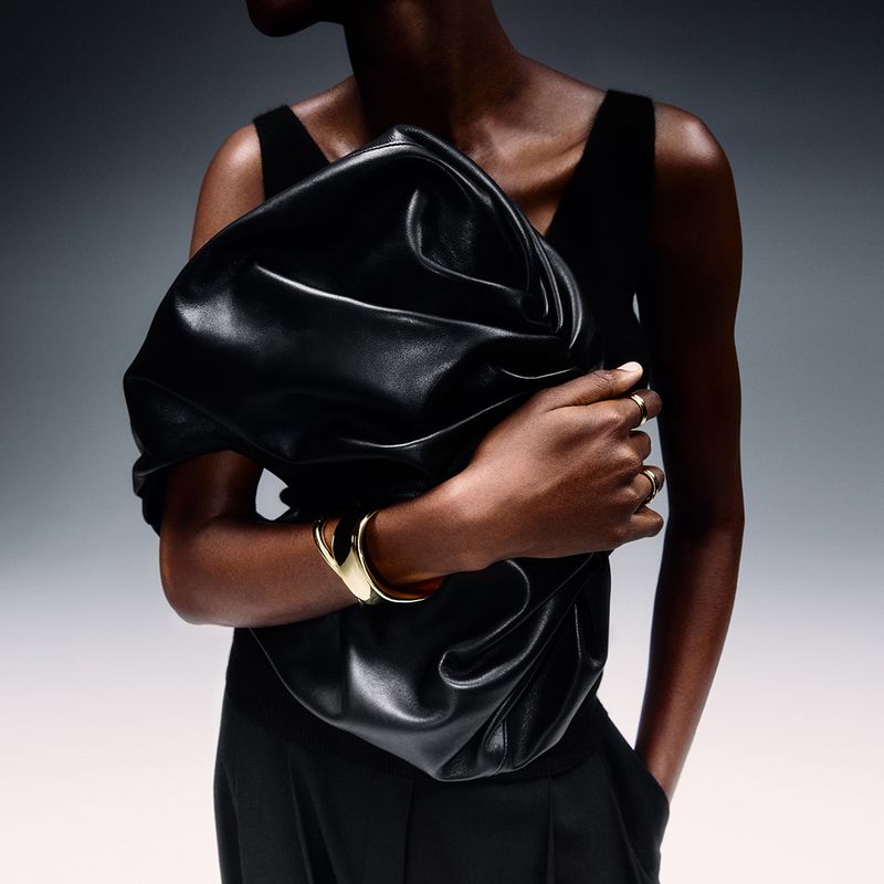 Woman in black top holding large black leather clutch bag and wearing gold jewelry against gray gradient background in studio shot.