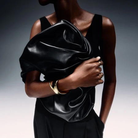Woman in black top holding large black leather clutch bag and wearing gold jewelry against gray gradient background in studio shot.