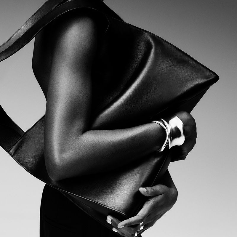 Black-and-white shot of a Black woman carrying a large black leather tote bag, wearing a silver cuff bracelet and rings.