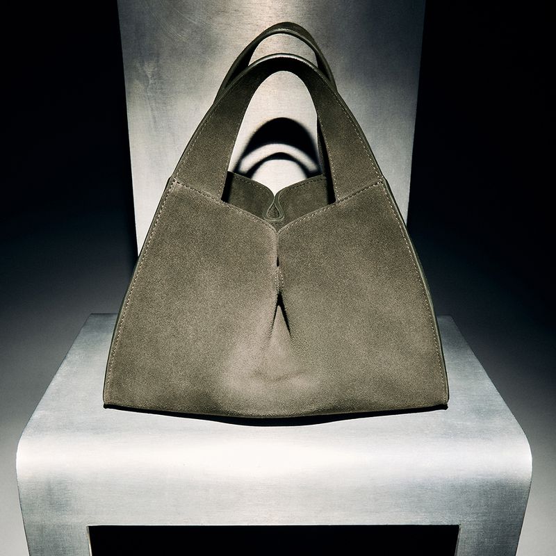 Elegant grey suede tote bag with double handles, displayed on a reflective silver stool against a dark background with soft studio lighting.