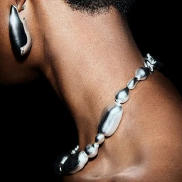 Close-up of a model wearing a chunky sculptural silver bead necklace and matching earring against a dark background.