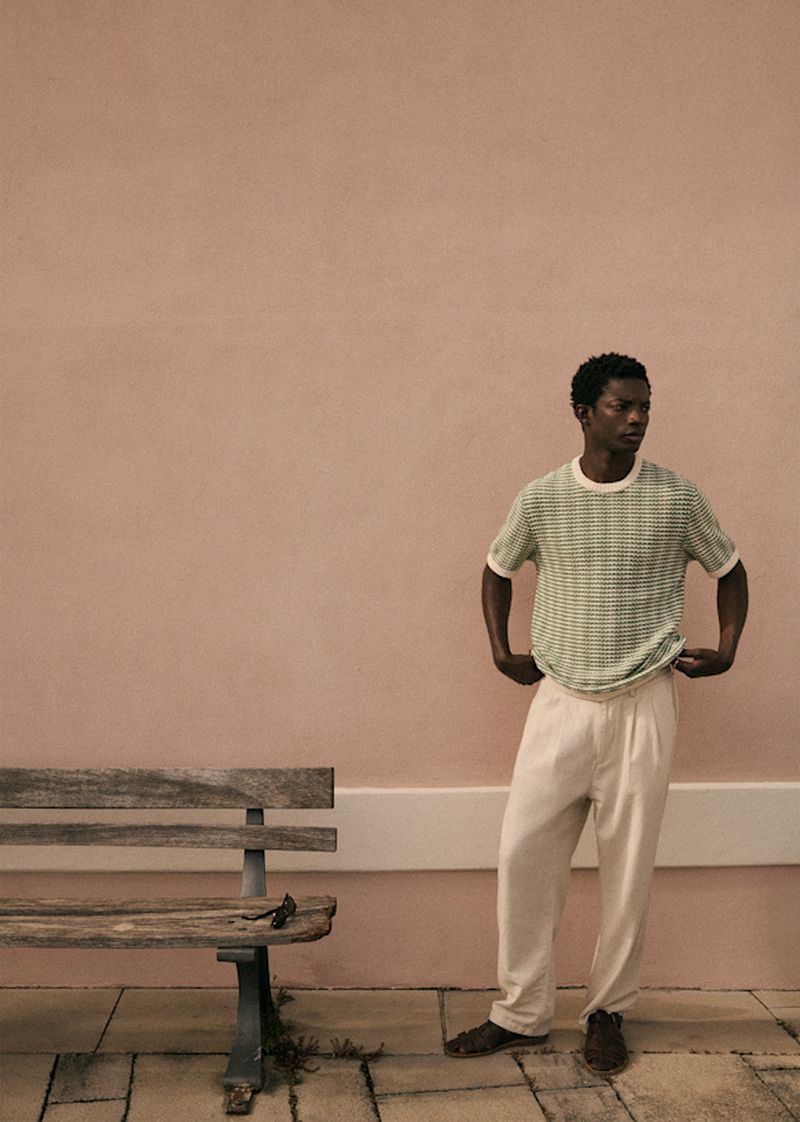 An adult Black man in a green patterned t-shirt and khaki pants standing outdoors next to a weathered wood bench against a pink wall.