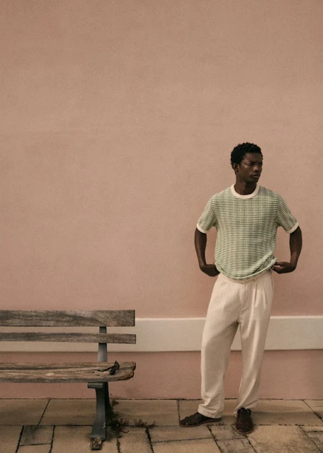 An adult Black man in a green patterned t-shirt and khaki pants standing outdoors next to a weathered wood bench against a pink wall.