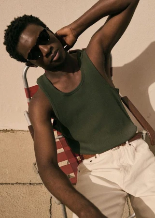 Young man in sunglasses and a green tank top, sitting on a lawn chair against a neutral wall, bathed in warm, diffuse light.