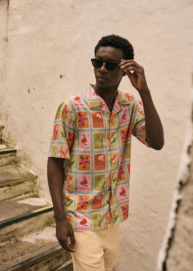 A dark-skinned man wears a floral-print shirt and sunglasses, standing on stairs outside in daylight. The shirt has red and yellow flowers.