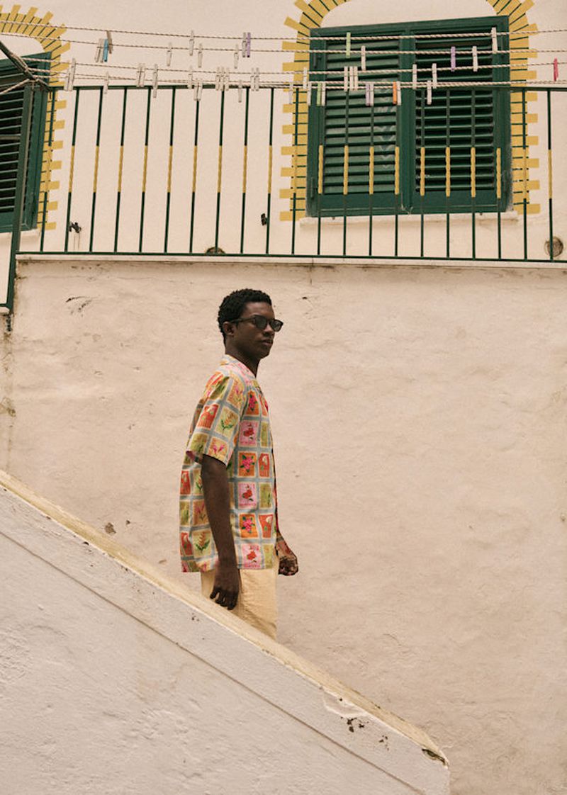 A man in sunglasses and a patterned shirt walking on stairs with a white building, green window, and clothesline in the background.