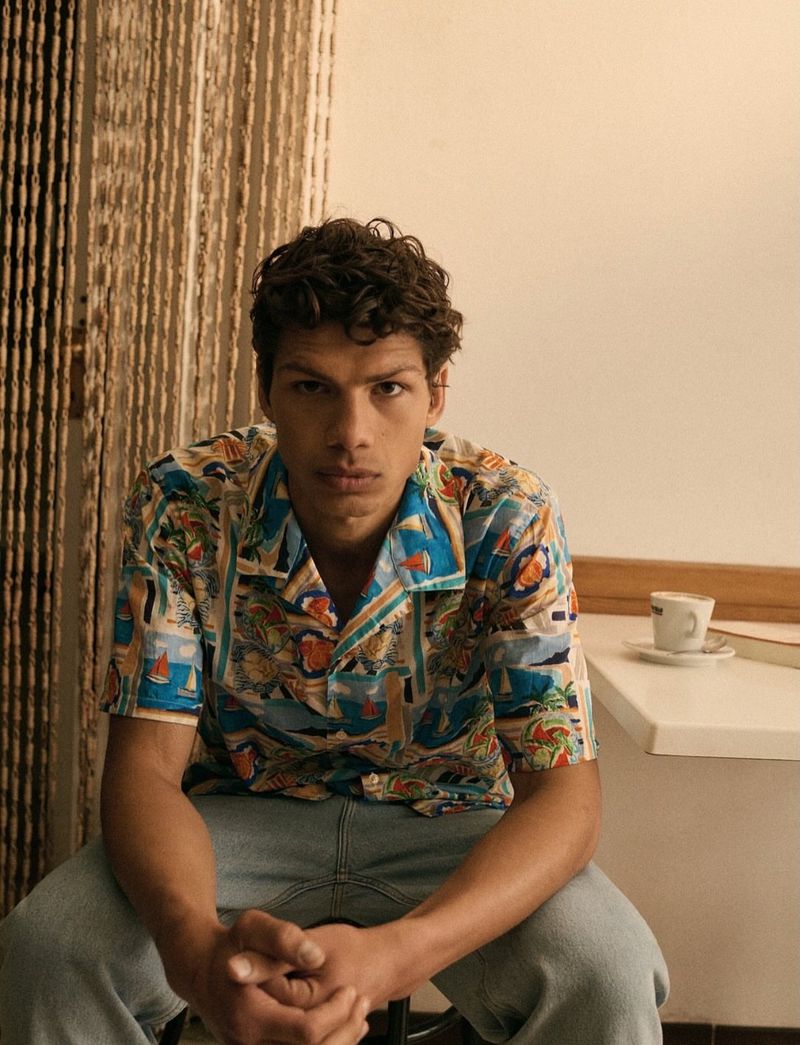 Adult male in a patterned shirt sitting on a chair indoors, with coffee and books on a table, soft light from a window.