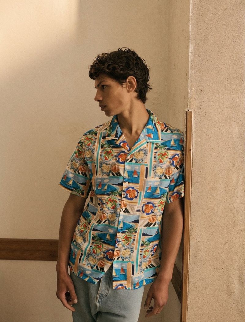 Male model in an open, short-sleeved patterned shirt with beach and vacation designs, posing against a corner wall indoors.