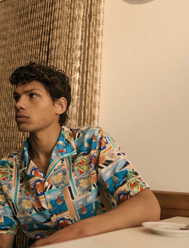 Young man wearing a patterned shirt sits at a white table in an indoor setting with a beaded curtain in the background.