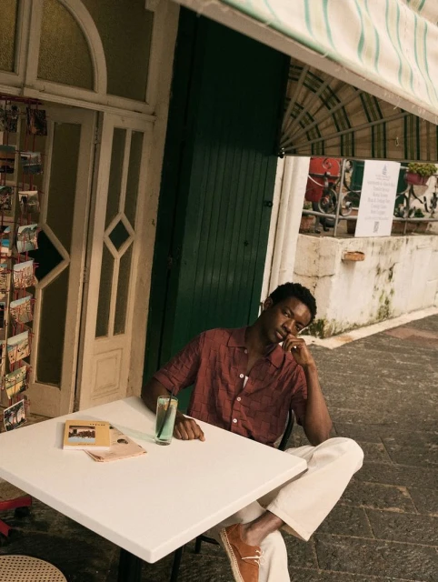 Black man in a button-down shirt and white pants seated at a white table on a patio with a drink and postcards.
