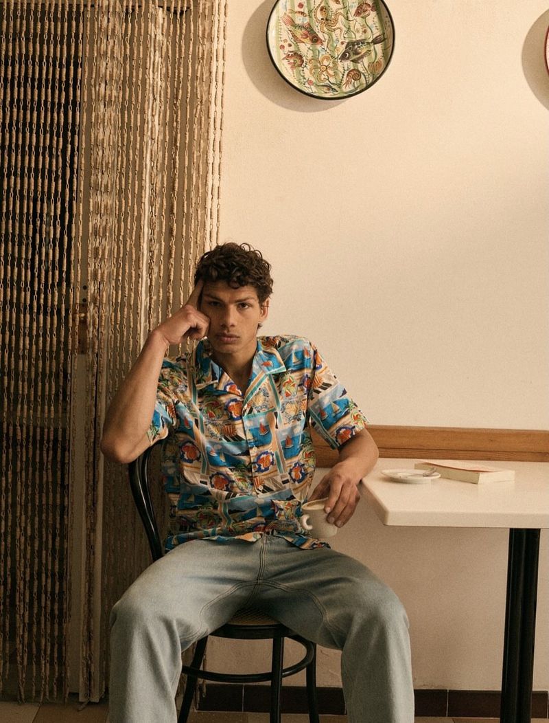 Portrait of a curly-haired male model in a patterned shirt sitting at a table with a coffee cup and book in an indoor setting.