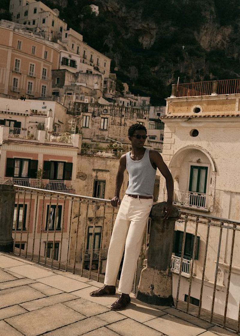 A man in a light blue tank top and white pants poses on a rooftop in an Italian coastal town, with buildings and mountains in the background.