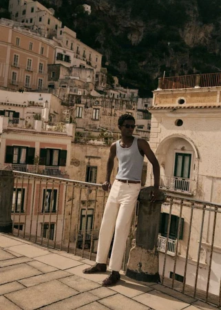 A man in a light blue tank top and white pants poses on a rooftop in an Italian coastal town, with buildings and mountains in the background.
