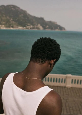 Back view of a Black man wearing a white tank top and a thin gold chain looking out at a turquoise ocean over a balustrade.