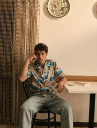 Man seated at a table wearing a printed short-sleeve shirt and light jeans while holding a coffee cup next to a beaded curtain.