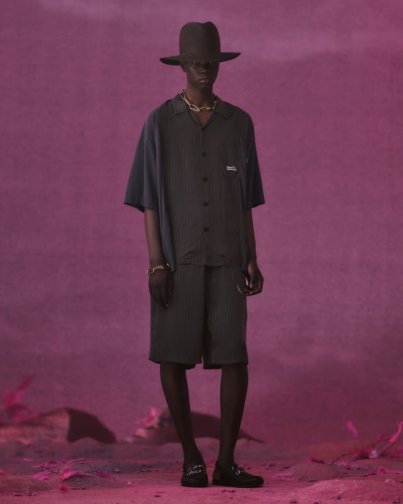 Full-length studio shot of a Black male model in a pinstripe short set, gold jewelry, and fedora hat against a magenta background.
