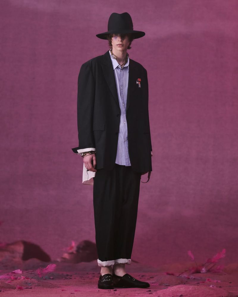 Full-length fashion shot of a person in a black hat, black blazer, light blue button-down shirt, and black pants standing in a pink environment.