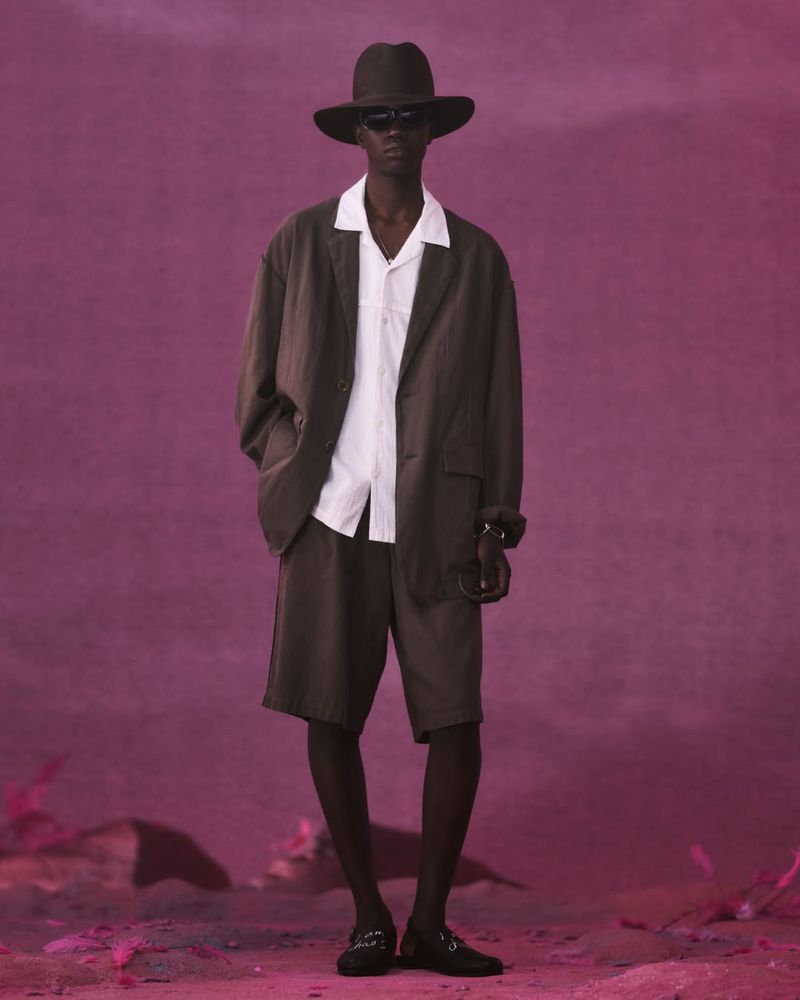 Model wearing a dark brown blazer, tailored shorts, white open-collar shirt, sunglasses, and wide-brim hat against a magenta set.