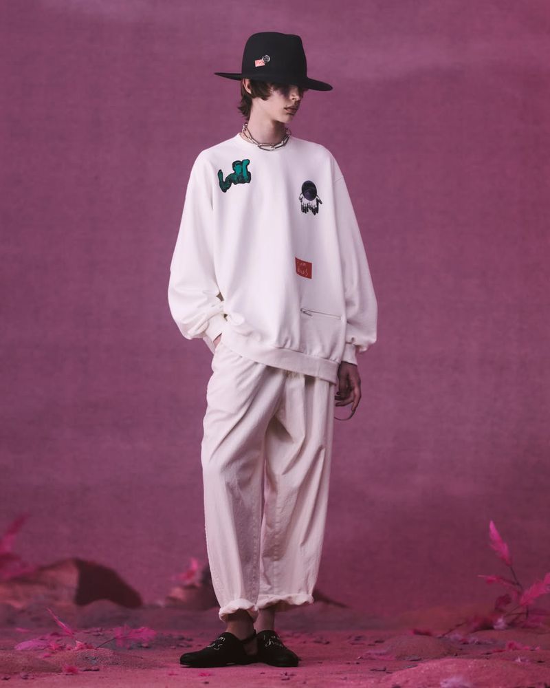 Full-length shot of a male model in a white sweatshirt with patches, loose cream pants, and black hat in a staged outdoor setting.