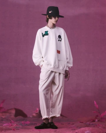 Full-length shot of a male model in a white sweatshirt with patches, loose cream pants, and black hat in a staged outdoor setting.