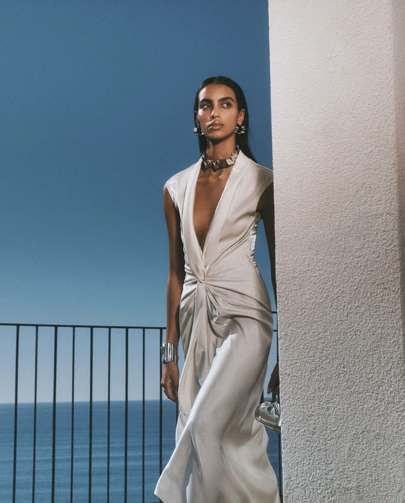 Elegant woman in sleeveless dress poses on a balcony against a blue sky and ocean backdrop, showcasing fashion and style with sophistication.