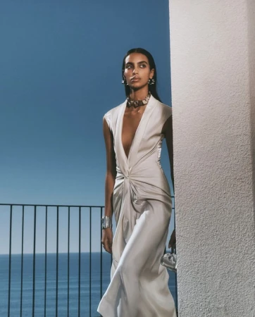 Elegant woman in sleeveless dress poses on a balcony against a blue sky and ocean backdrop, showcasing fashion and style with sophistication.