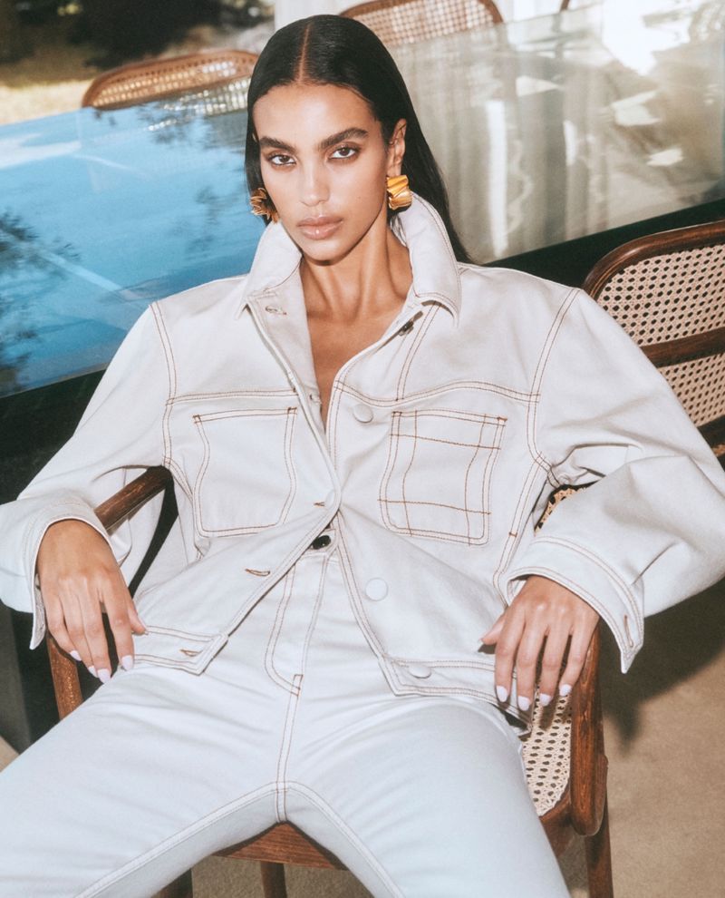 Elegant woman in a cream-colored denim jacket and jeans sitting in a rattan chair; stylish gold earrings complete the fashionable ensemble.