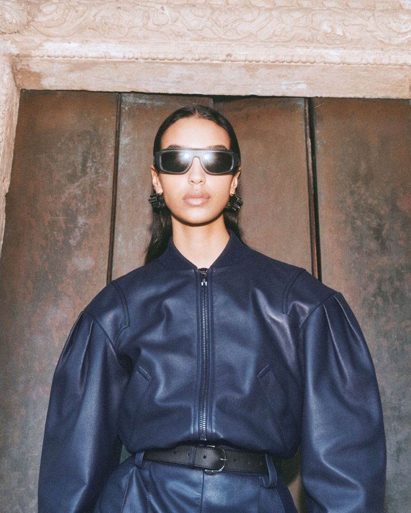 Stylish woman with sunglasses poses in a blue leather jacket and pants in front of a textured wall, exuding modern fashion.