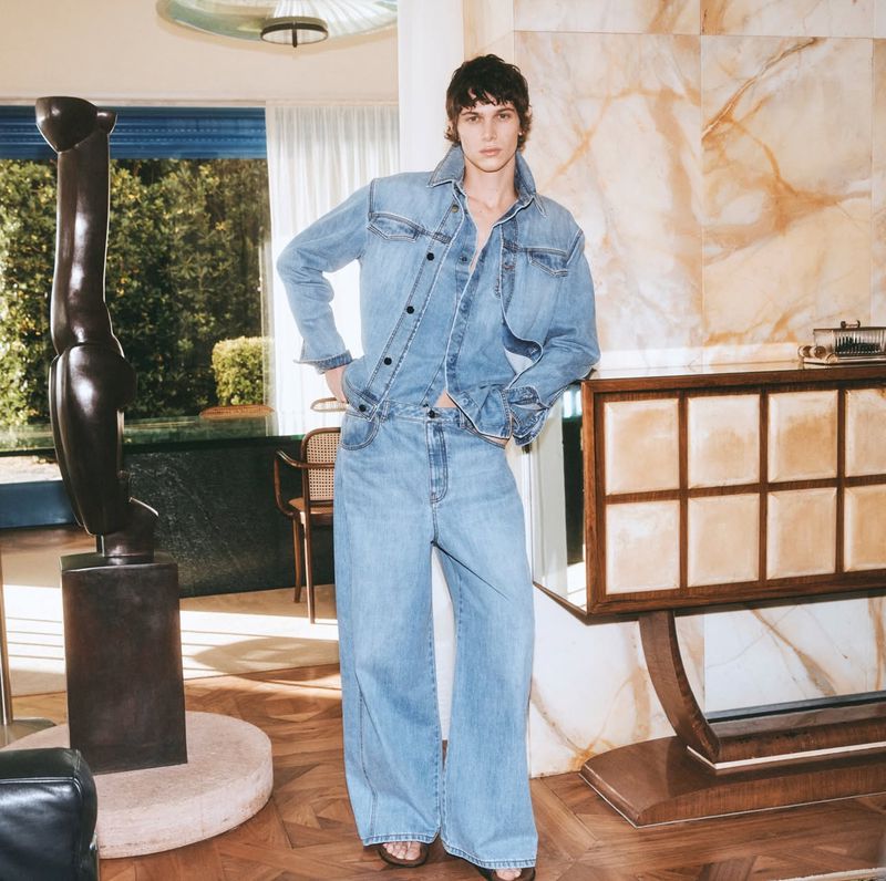 An adult male fashion model wearing a denim jacket and jeans outfit poses in an interior setting with sculptures and furniture.