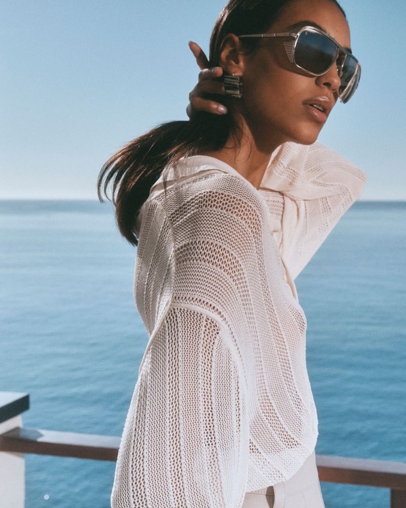 Woman with sunglasses and earrings poses in a knitted sweater with an ocean and sky backdrop, capturing a blend of fashion and nature.