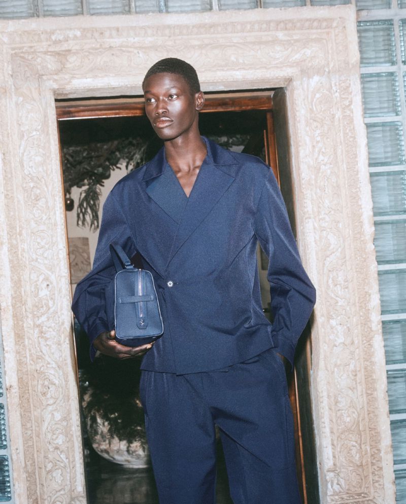 A dark-skinned male model wearing a navy blue suit and carrying a matching bag poses in a doorway with ornate plaster details.